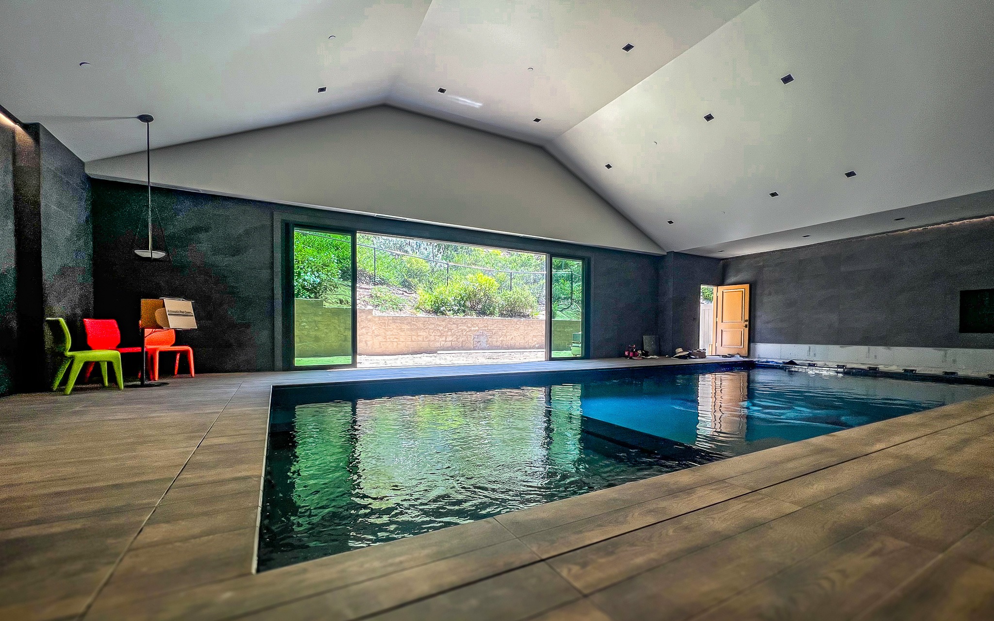Modern indoor swimming pool with dark gray walls and large windows overlooking a garden. there's a colorful seating area near the windows.