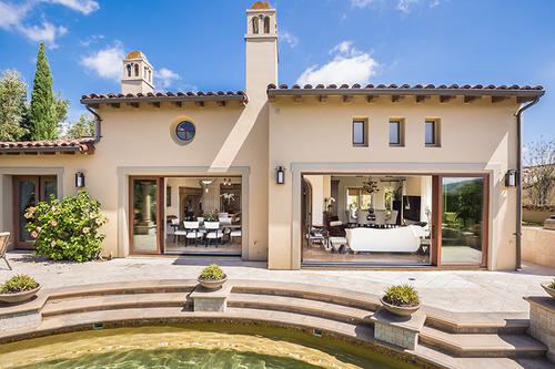 Single-story house with beige stucco exterior, arched windows, and double glass doors opening to a patio with steps leading to a circular pool. The sky is clear and the surroundings include plants.