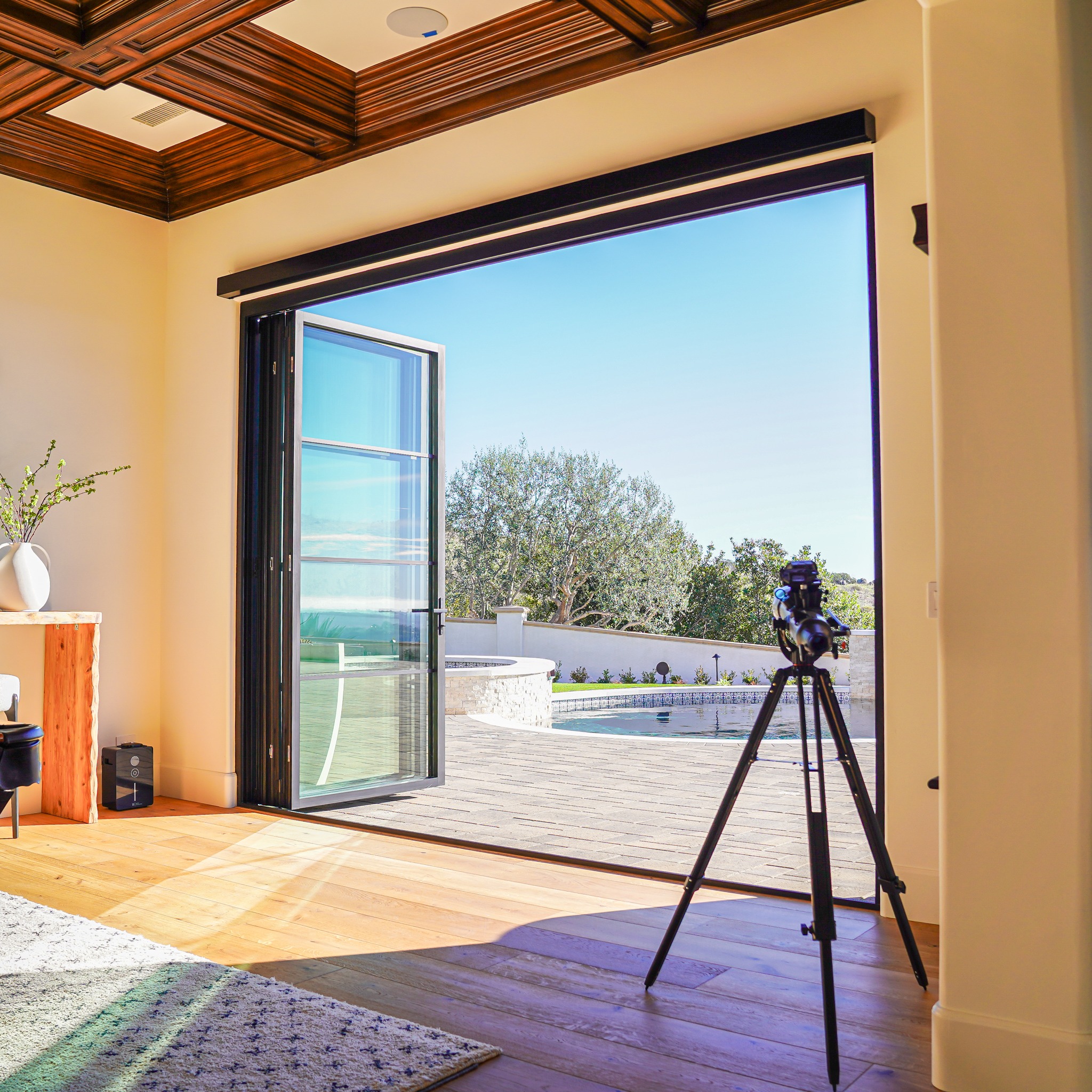 A camera on a tripod is set up at an open patio door, overlooking a terrace with a pool and trees beyond, shot from inside a modern room with wooden floors and ceiling.