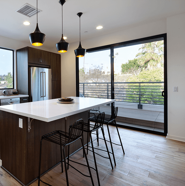 A modern kitchen with a central island, three bar stools, pendant lights, stainless steel appliances, and large sliding glass doors leading to a balcony with a view of trees.