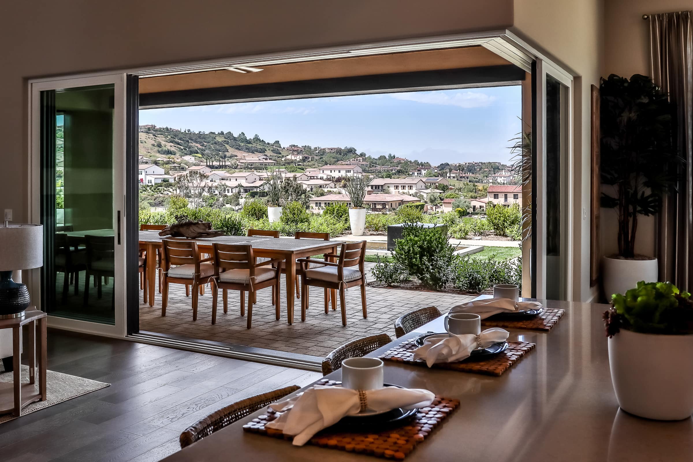 Open sliding glass doors reveal an outdoor patio with a dining set overlooking a suburban neighborhood, seen from a modern indoor kitchen with a breakfast bar and place settings.