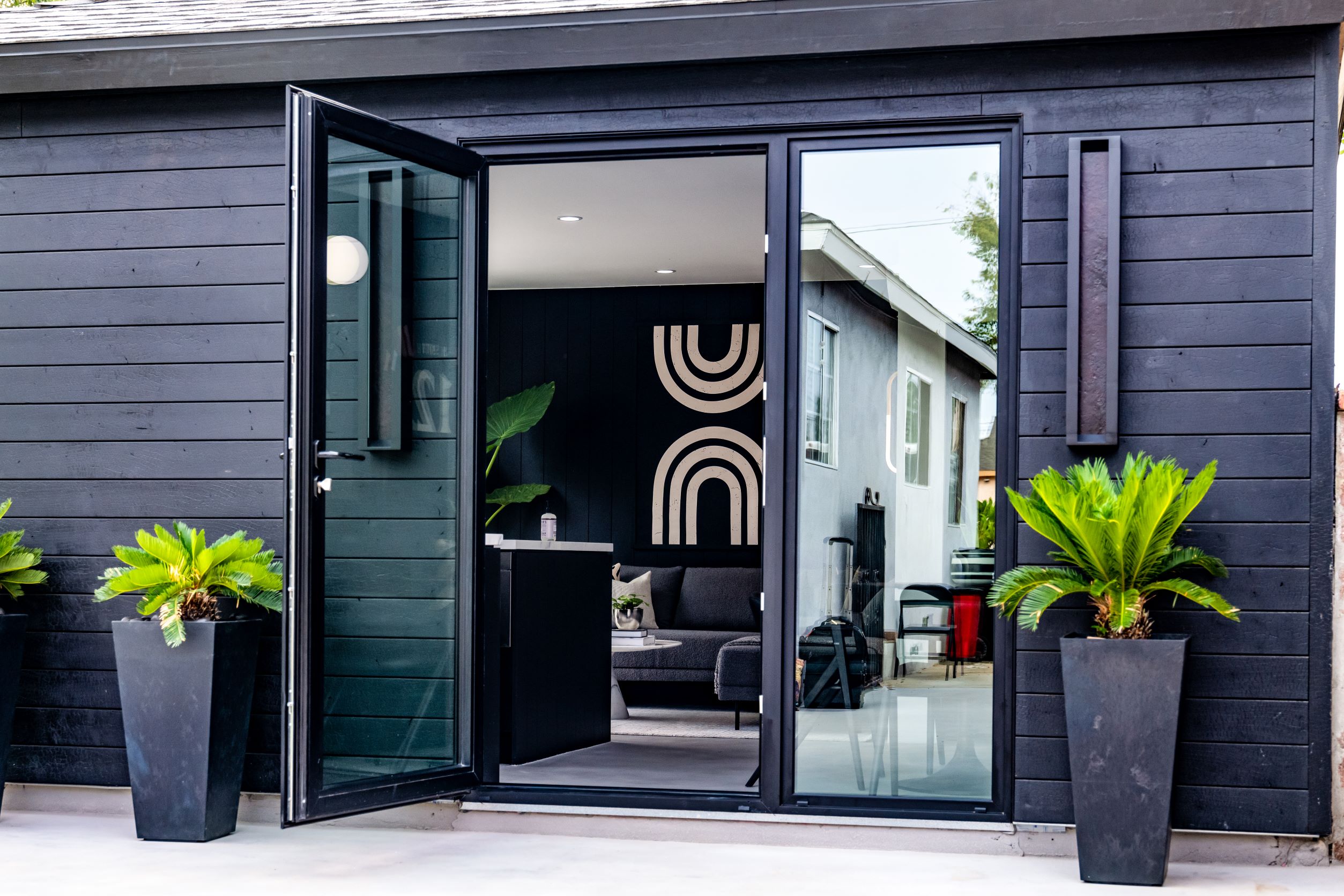 A modern black building with an open glass door, large potted plants on either side, and a glimpse of a minimalist interior with abstract wall art.